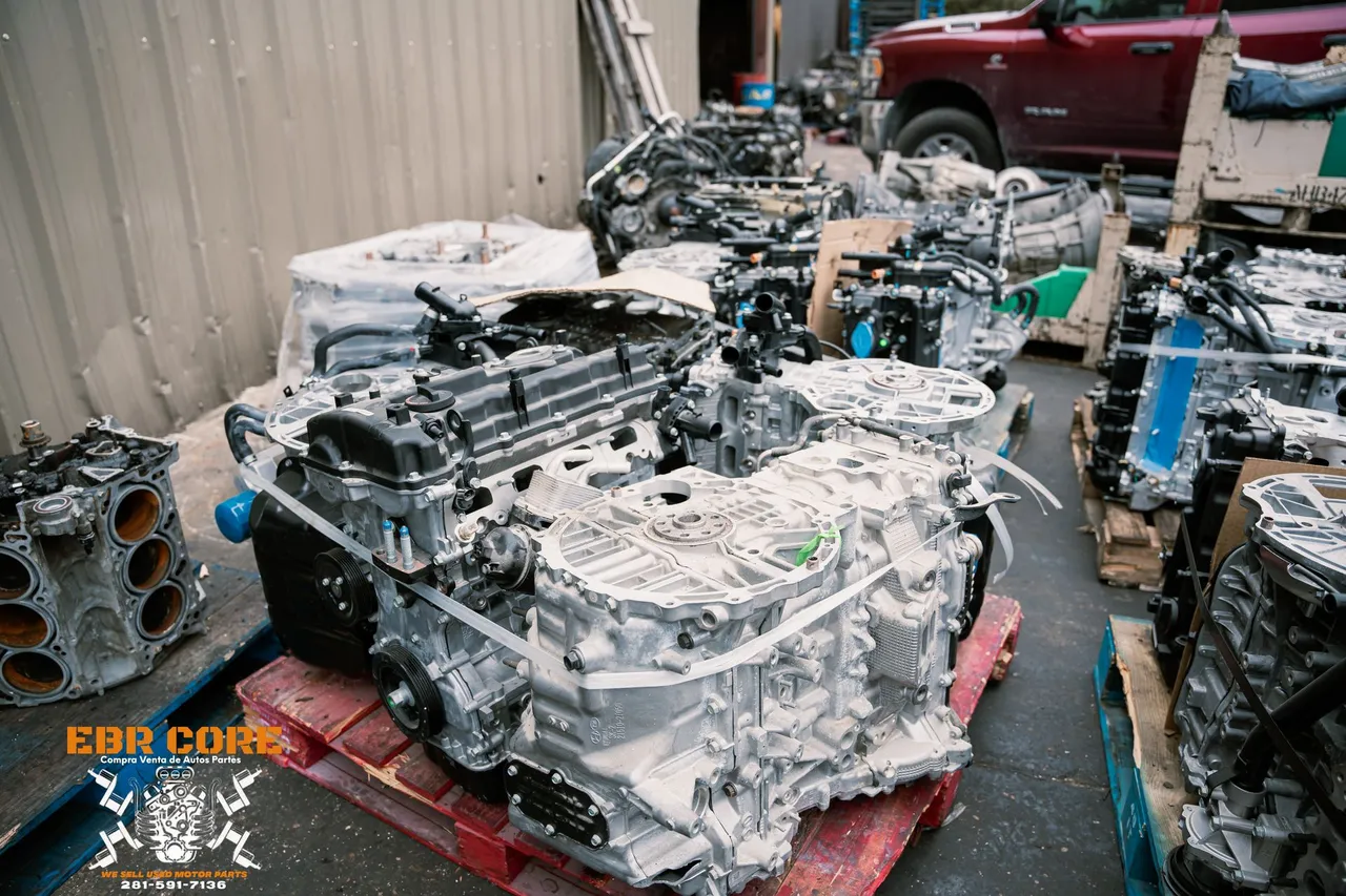 Engines piled on pallets in a junkyard, covered in a white substance. A red truck is in the background.