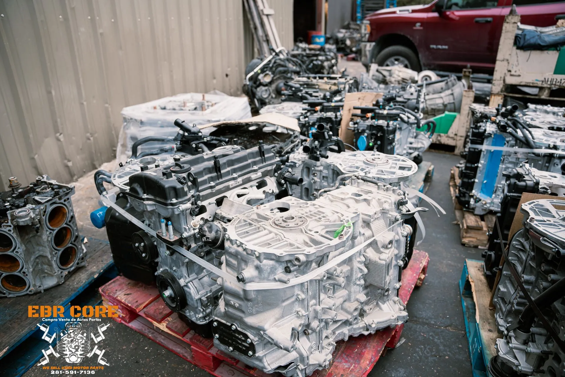 Engines piled on pallets in a junkyard, covered in a white substance. A red truck is in the background.